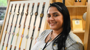 Fotografía de Melany Bolívar sonriendo y al fondo, se ven los relojes artesanales que elabora en su taller